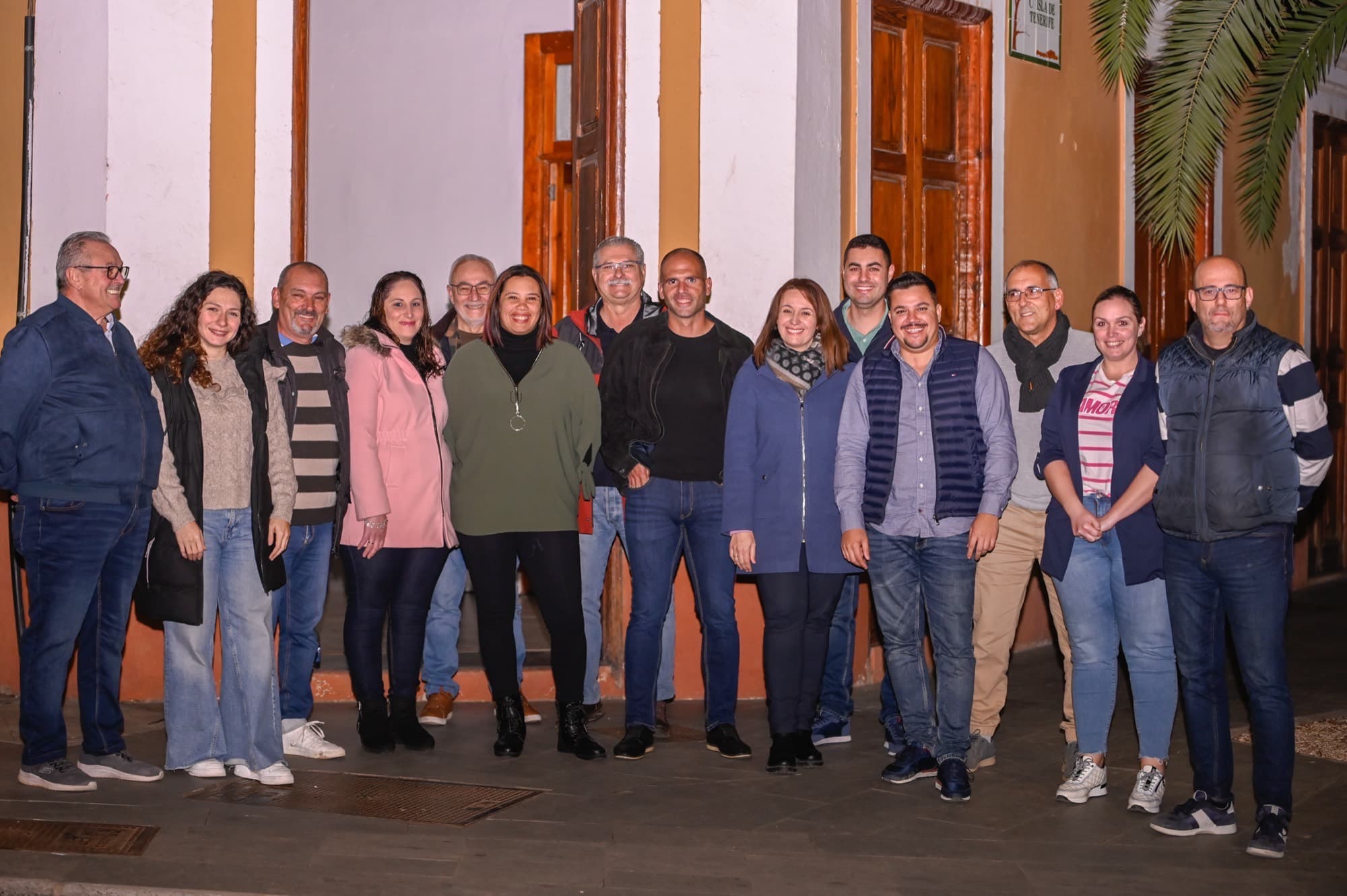 Grupo de una quincena de personas posando frente a un edificio con puertas de madera y una palmera al lado.